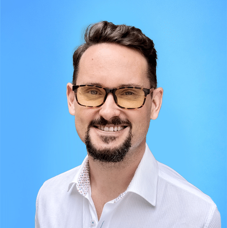A man with short brown hair, a goatee, and glasses smiles at the camera while wearing a white collared shirt against a blue background.