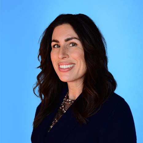 Woman with long dark hair wearing a dark blazer and patterned top, smiling, posed in front of a plain blue background.