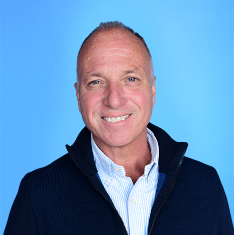 A middle-aged man with short hair smiles at the camera, wearing a navy jacket and light striped shirt, against a blue background.