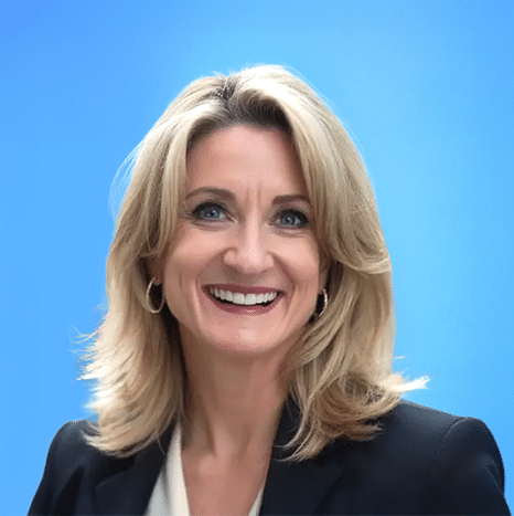 A woman with shoulder-length blonde hair, wearing a black blazer and white top, smiles in front of a plain blue background.