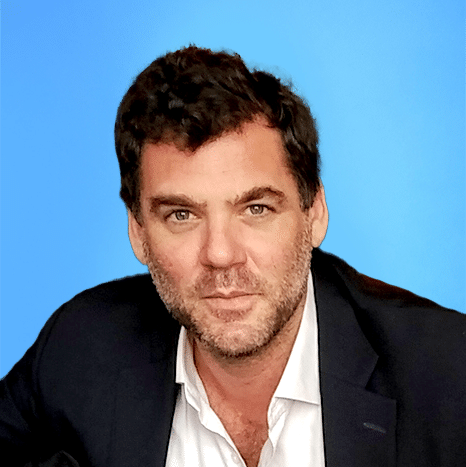 A man with short dark hair and a beard wears a white shirt and dark blazer, seated in front of a solid blue background.