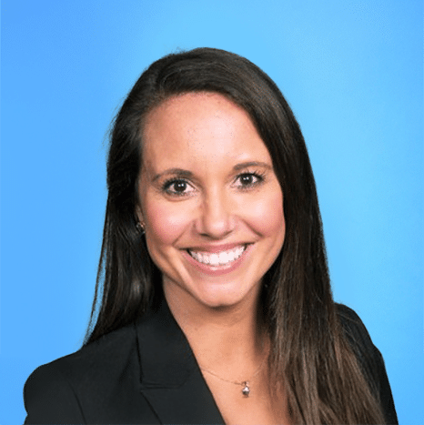 Woman with long brown hair wearing a black blazer smiles at the camera against a solid light blue background.
