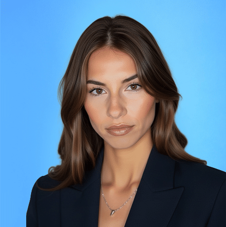 Woman with long brown hair wearing a dark blazer and a necklace poses against a plain blue background.