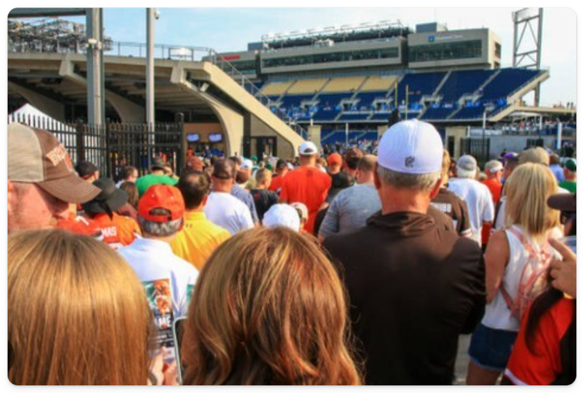 A large crowd of people, many wearing hats and sports jerseys, stands outside a stadium entrance on a sunny day.