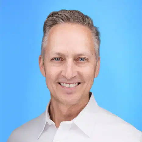 Smiling middle-aged man with short gray hair wearing a white collared shirt, posed against a plain blue background.