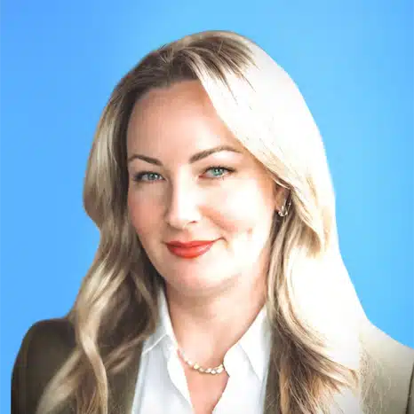 Professional woman with long blonde hair, wearing a white blouse, dark blazer, and pearl necklace, smiling against a plain blue background.