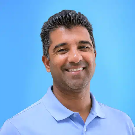 A man with short dark hair and light stubble, wearing a light blue collared shirt, smiles in front of a plain blue background.