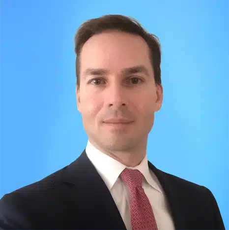 A man in a dark suit, white shirt, and pink tie poses against a plain blue background.