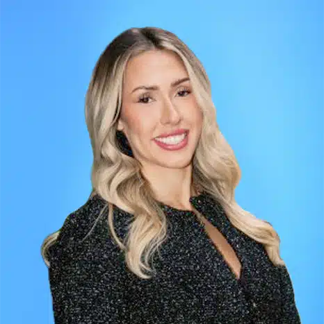 Woman with long blonde hair wearing a dark textured jacket, smiling in front of a plain blue background.