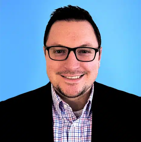 Man wearing glasses, a checked shirt, and dark blazer, smiling in front of a plain blue background.