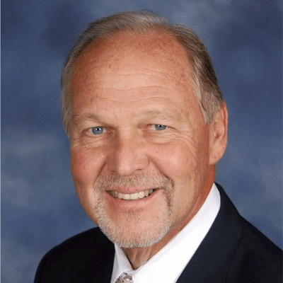Smiling older man with light skin, short light hair, and a trimmed beard, wearing a dark suit, white shirt, and tie, posing in front of a blue background.